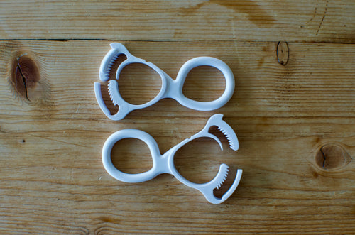 Two white Antenna Mate clips laid on a weathered wood surface, each with a figure-eight body: a smooth ring at one end for threading webbing and a toothed jaw at the other that grips the antenna.