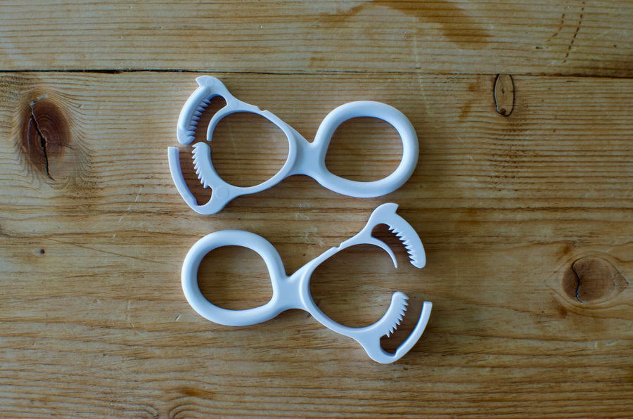 Two white Antenna Mate clips laid on a weathered wood surface, each with a figure-eight body: a smooth ring at one end for threading webbing and a toothed jaw at the other that grips the antenna.