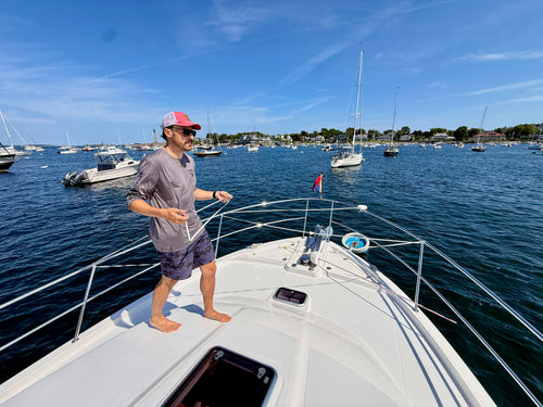A boater standing on the foredeck pulls LINEGUARD's white webbing across the bow rails of a powerboat moored in a New England harbor, showing the deterrent being deployed by hand.
